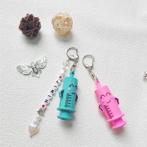 Puede incluir: Una colección de llaveros y objetos decorativos. Dos llaveros presentan dijes de jeringa sonrientes en verde azulado y rosa. Un dije de cuentas deletrea "JIMIN". Otros artículos incluyen un colgante de mariposa plateada y dos bolas decorativas.