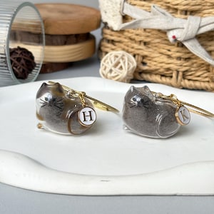 May include: Two cat-shaped keychains. One is brown and tan, the other is gray. Each keychain has a gold-colored metal wire and a small, round charm with a letter. The brown cat has an "H" charm, and the gray cat has a "K" charm.