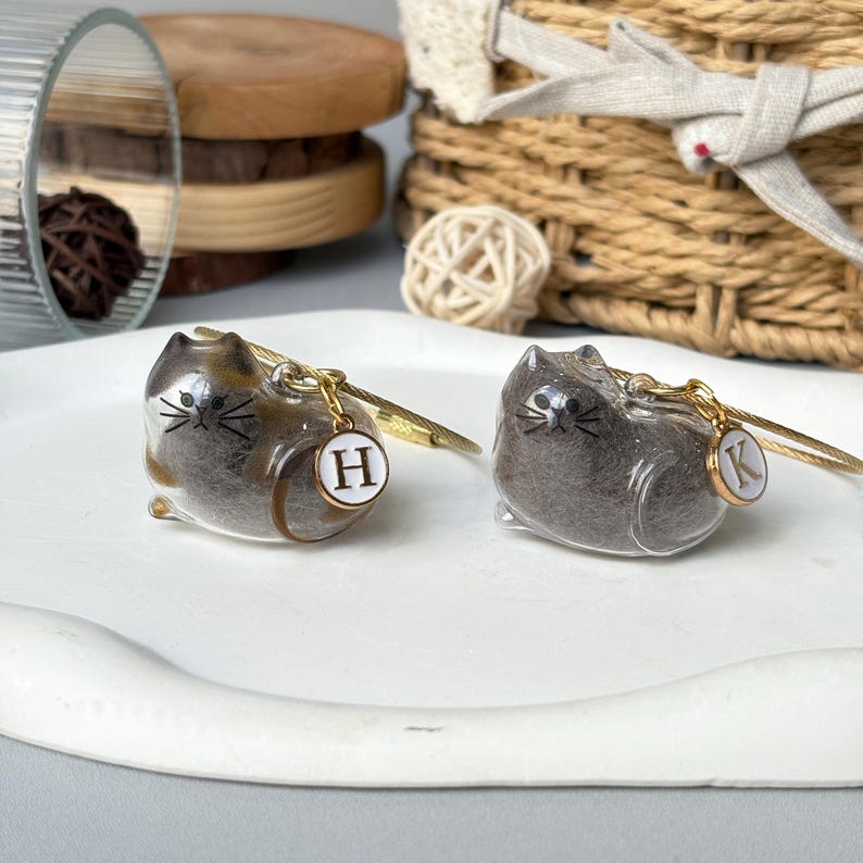 May include: Two cat-shaped keychains. One is brown and tan, the other is gray. Each keychain has a gold-colored metal wire and a small, round charm with a letter. The brown cat has an "H" charm, and the gray cat has a "K" charm.