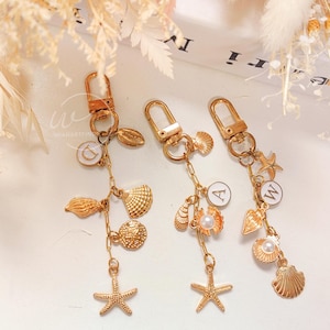 May include: Three gold keychains with charms. Each keychain has a different letter charm, "D", "A", and "W". The charms include seashells, starfish, and a pearl.