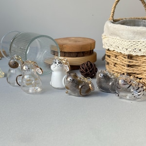 May include: A collection of glass cat figurines with gold-colored accents. Some cats are clear, others are white or brown. Each cat has a small gold charm. A woven basket and wooden coasters are in the background.