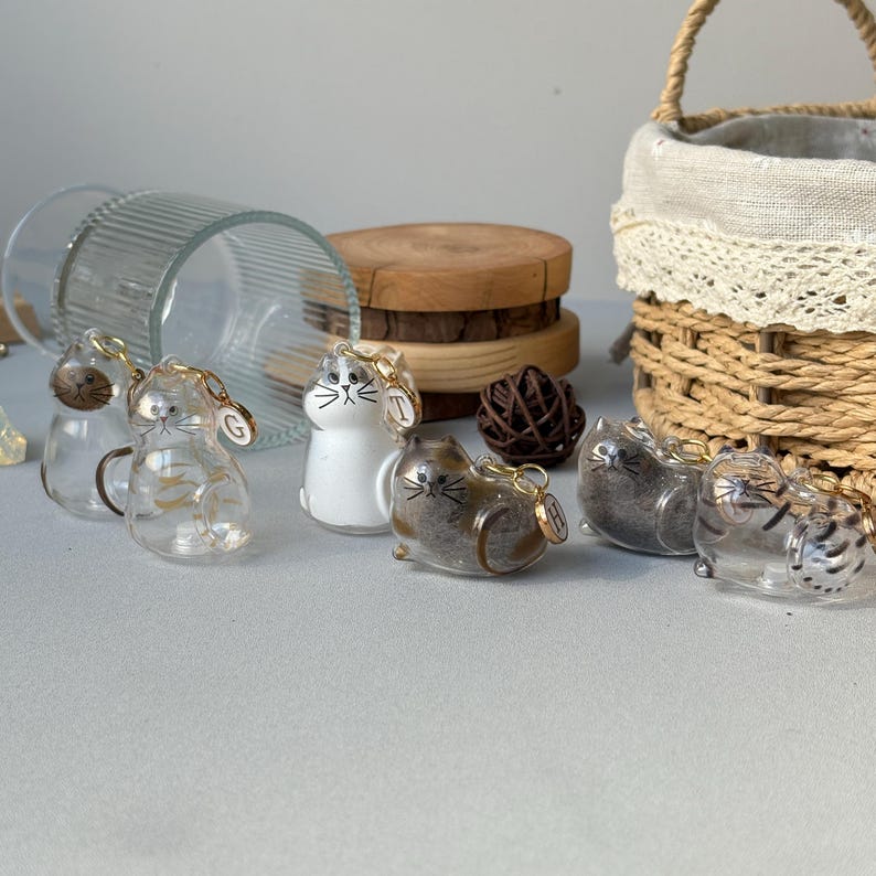 May include: A collection of glass cat figurines with gold-colored accents. Some cats are clear, others are white or brown. Each cat has a small gold charm. A woven basket and wooden coasters are in the background.