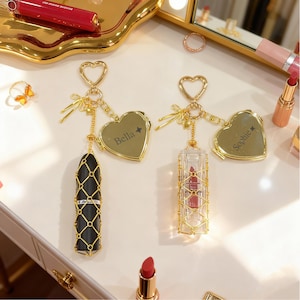 Könnte beinhalten: Zwei Lippenstift-Schlüsselanhänger mit goldfarbenen herzförmigen Anhängern. Einer hat ein schwarzes Lippenstiftgehäuse, der andere ein klares Gehäuse. Beide haben goldfarbene Akzente und die Namen "Bella" und "Sophie" auf den Anhängern eingraviert. Im Hintergrund befinden sich weitere Make-up-Artikel.