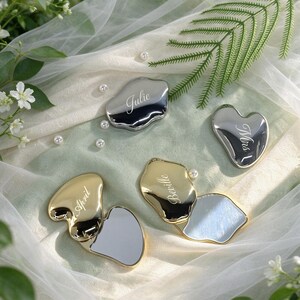 May include: Four irregular, heart-shaped compact mirrors with metallic finishes in silver and gold. Two mirrors are closed, displaying the engraved names Julie and Mrs. Two mirrors are open, showing the reflective surface and the names April and Beville.