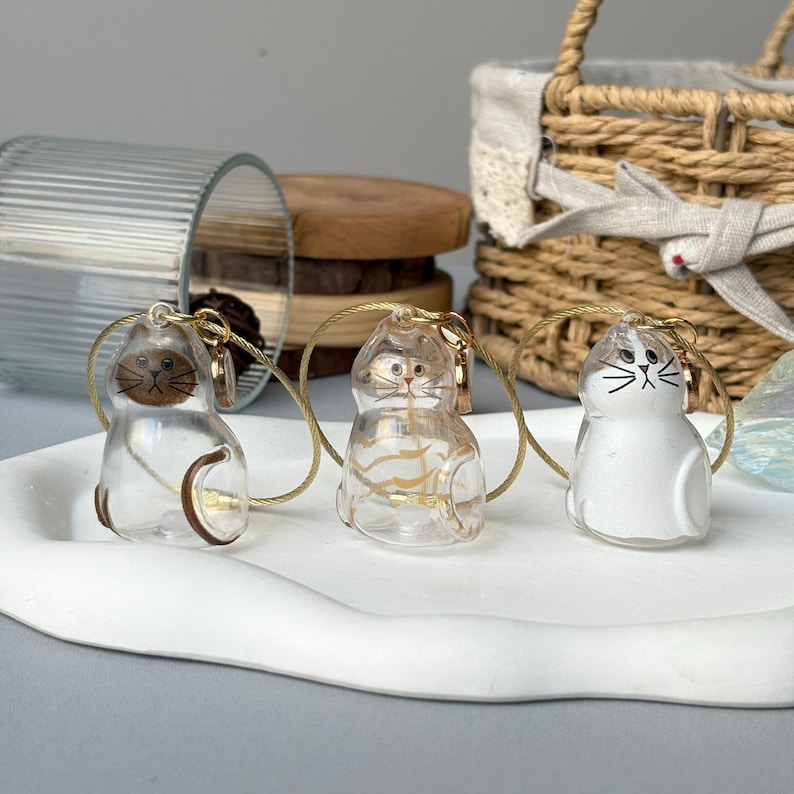 May include: Three clear cat-shaped keychains with gold-colored accents. The keychains are on a white tray. One cat is brown, one has gold stripes, and the third is white with black markings. Each keychain has a gold-colored wire loop.