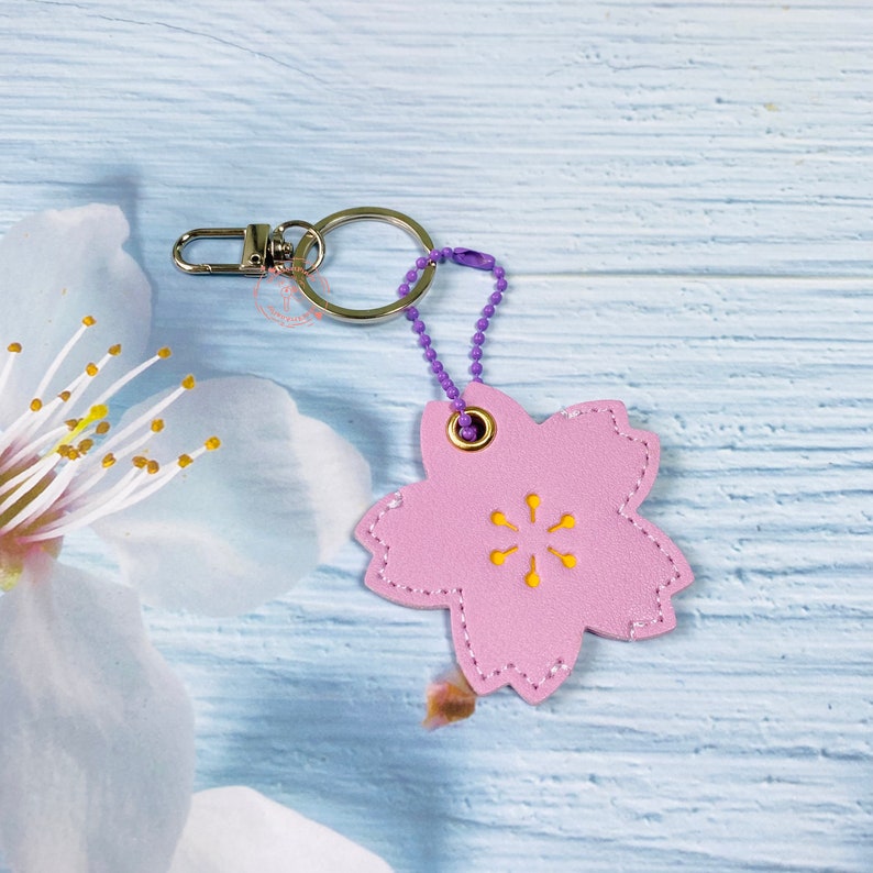 May include: Pink leather keychain shaped like a cherry blossom with a yellow center and a silver ring. The keychain is attached to a purple chain.