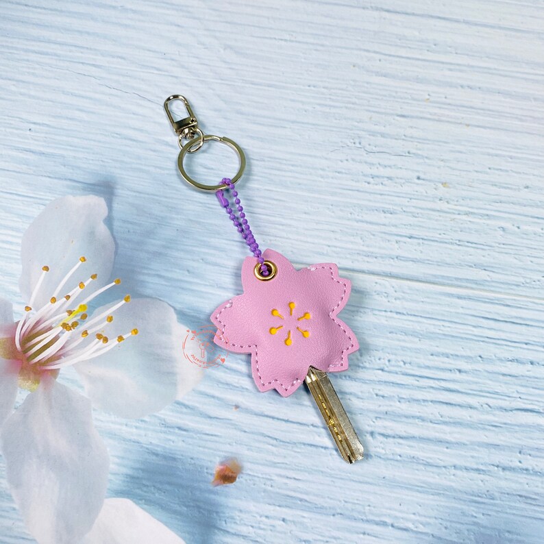 May include: A pink leather keychain shaped like a cherry blossom with a yellow center. The keychain has a silver ring and a purple chain.