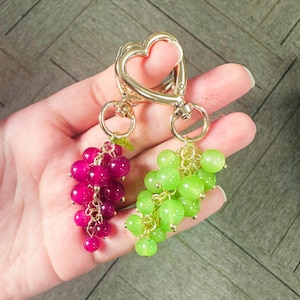 May include: A gold heart-shaped keychain with two charms: a bunch of pink grapes and a bunch of green grapes.