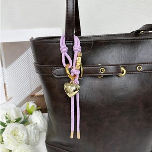 May include: A dark brown tote bag with a light purple charm. The charm features a gold heart pendant and the name "KATE" in gold letters. The bag has a buckle detail and gold hardware.