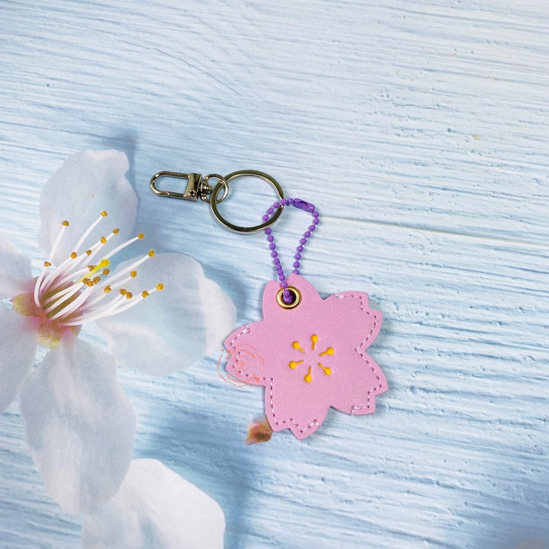 May include: A pink leather keychain with a cherry blossom design. The keychain has a purple chain and a silver ring.