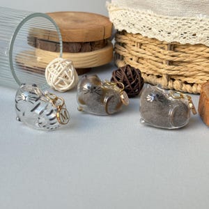 May include: Three clear cat-shaped keychains with gold-colored accents. One keychain has a silver striped design, and another has a gold initial charm. The keychains are on a light gray surface, with a glass, wooden slices, and a woven basket in the background.