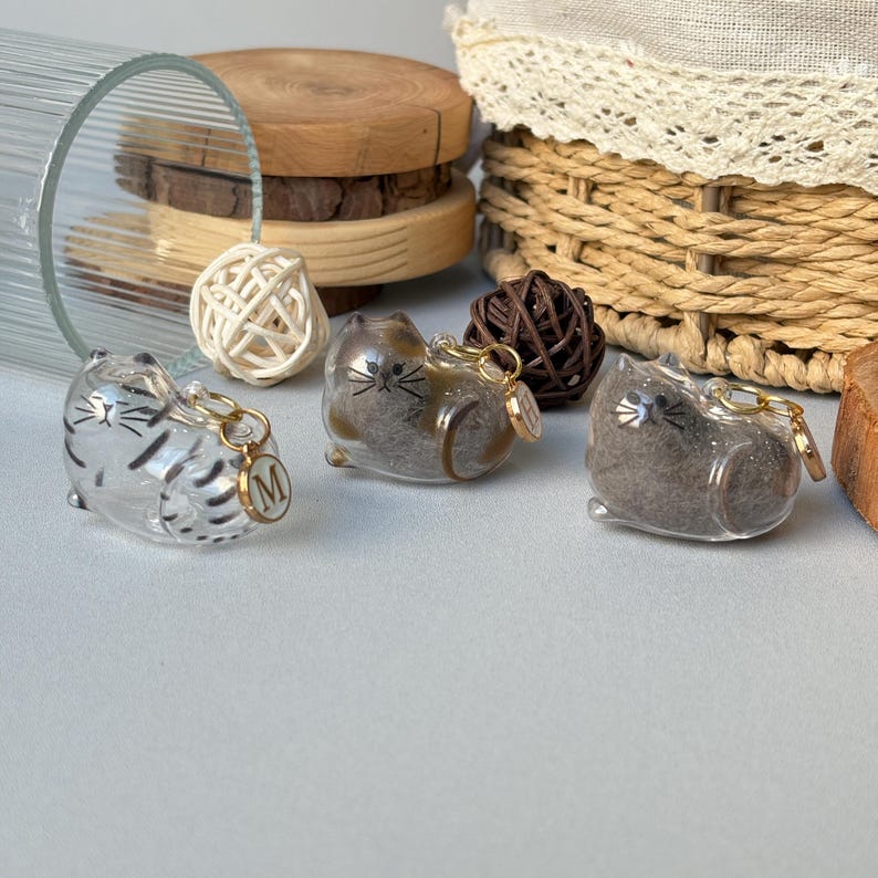 May include: Three clear cat-shaped keychains with gold-colored accents. One keychain has a silver striped design, and another has a gold initial charm. The keychains are on a light gray surface, with a glass, wooden slices, and a woven basket in the background.