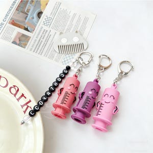 May include: Three syringe-shaped keychains in shades of pink and purple, each with a smiling face and measurement markings. One keychain has a beaded strand with the letters "SCCOYATHBOS". The keychains are attached to silver keyrings.