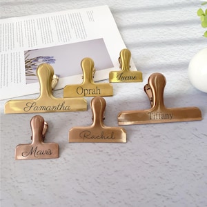 May include: Six metal binder clips in gold and copper tones. Each clip is engraved with a name: Samantha, Oprah, Lucine, Tiffany, Rachel, and Mavis. The clips are displayed on a light surface, with a magazine page in the background.