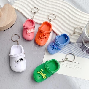 May include: Small clog-shaped keyrings in pink, orange, blue, white, and green. Each keyring features a name: Stella, Jeff, Dylan, Poppy, and Judy. The keyrings are attached to silver rings.