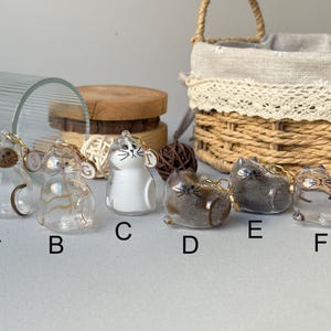 May include: A collection of six glass cat figurines with gold keychains. The cats are in various colors and patterns, including clear, white, brown, and gray. Each cat has a unique design, with some featuring stripes or spots. A basket and wooden slices are in the background.
