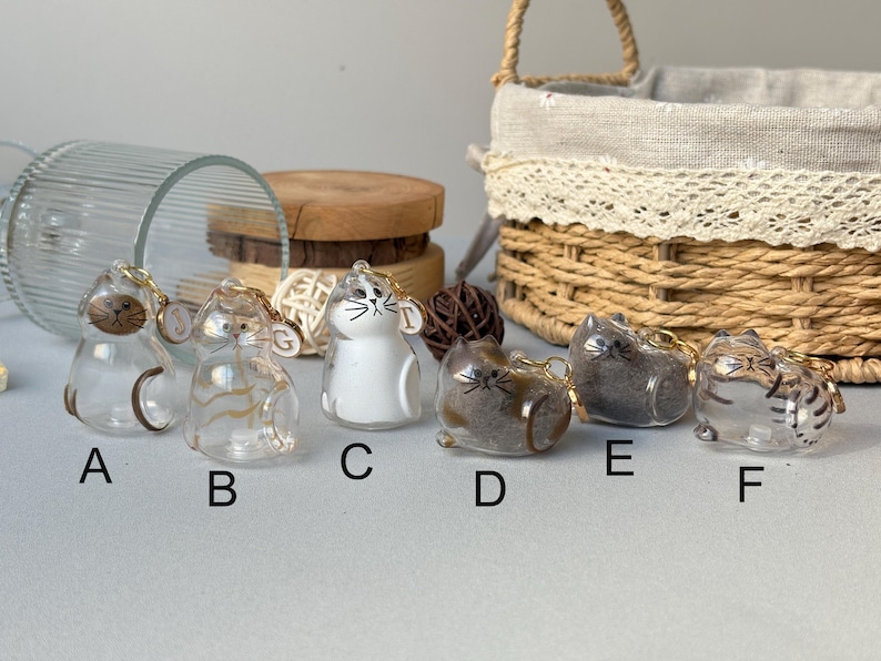 May include: A collection of six glass cat figurines with gold keychains. The cats are in various colors and patterns, including clear, white, brown, and gray. Each cat has a unique design, with some featuring stripes or spots. A basket and wooden slices are in the background.