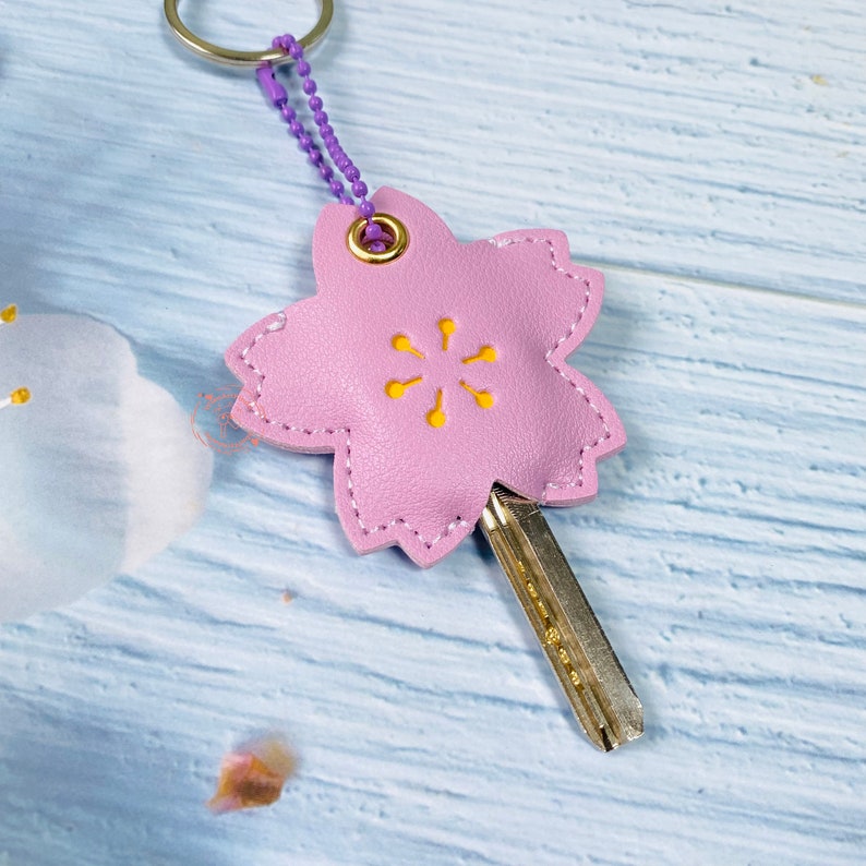 May include: A pink leather keychain shaped like a cherry blossom with a yellow flower design and a metal key ring. The keychain is attached to a silver key.