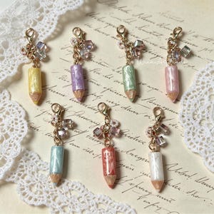 May include: Seven pencil-shaped charms in pastel shades of yellow, purple, green, pink, blue, red, and white. Each charm features a gold-coloured clasp and is decorated with a cluster of beads and crystals. The pencils have a gold-coloured tip.