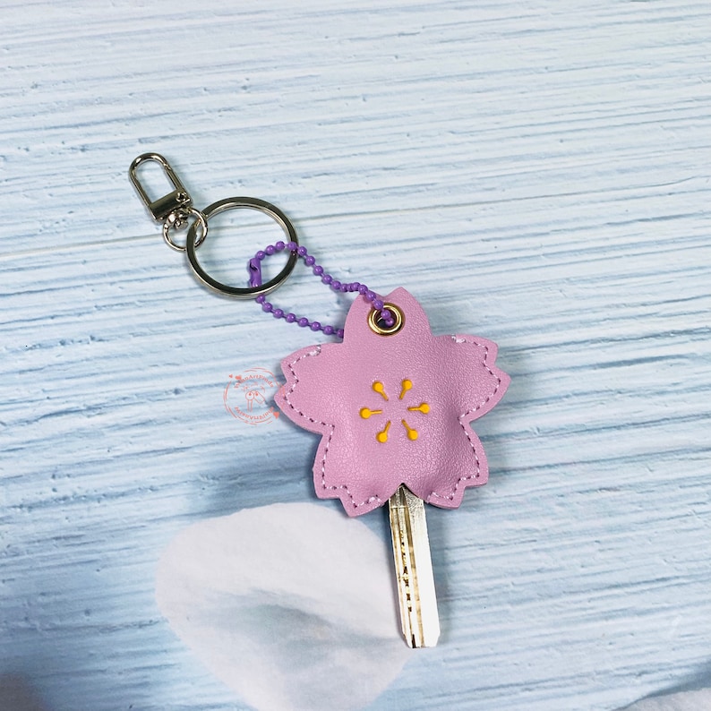 May include: A pink leather key cover shaped like a cherry blossom with a yellow center. The key cover is attached to a silver key ring with a purple chain.