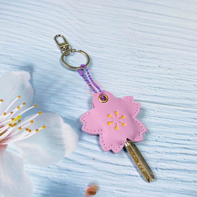 May include: A pink leather keychain shaped like a cherry blossom with a yellow center. The keychain has a silver ring and a purple chain.