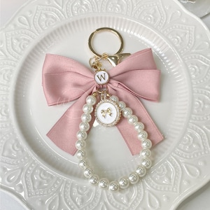 Può includere: Un fiocco di raso rosa con un portachiavi a collana di perle. Il portachiavi presenta un ciondolo dorato con un fiocco in smalto bianco e la lettera "W" in oro.