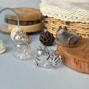 May include: Three clear cat-shaped keychains with gold-colored hardware. One keychain has gold leaf accents, and another has black stripes. The third keychain is a brown and clear cat. The keychains are on a light blue surface.