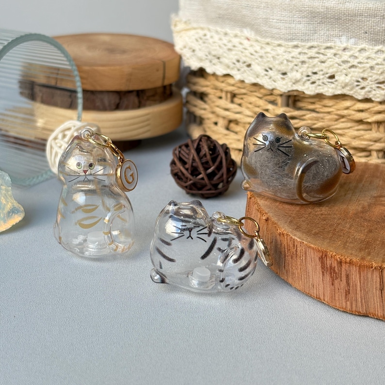 May include: Three clear cat-shaped keychains with gold-colored hardware. One keychain has gold leaf accents, and another has black stripes. The third keychain is a brown and clear cat. The keychains are on a light blue surface.