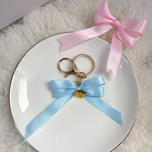 Può includere: Un portachiavi in metallo dorato con un fiocco in nastro di raso azzurro e un piccolo ciondolo circolare dorato inciso con la lettera V. Un fiocco in nastro di raso rosa chiaro separato è posizionato sopra il portachiavi su un piatto bianco.