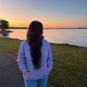 May include: A person wearing a lavender hoodie with the word "becoming" embroidered on the chest, paired with light blue jeans. The person is standing on a paved path near a grassy area, with a lake and sunset in the background.
