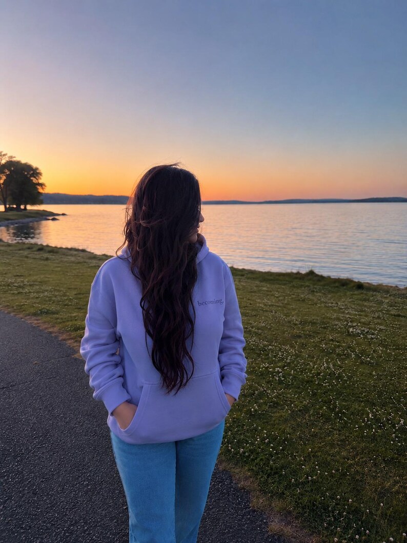 May include: A person wearing a lavender hoodie with the word "becoming" embroidered on the chest, paired with light blue jeans. The person is standing on a paved path near a grassy area, with a lake and sunset in the background.