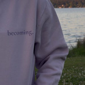May include: A lavender hoodie with the word "becoming" printed on the front. The hoodie has a drawstring hood and a kangaroo pocket. The background shows a body of water and a treeline.