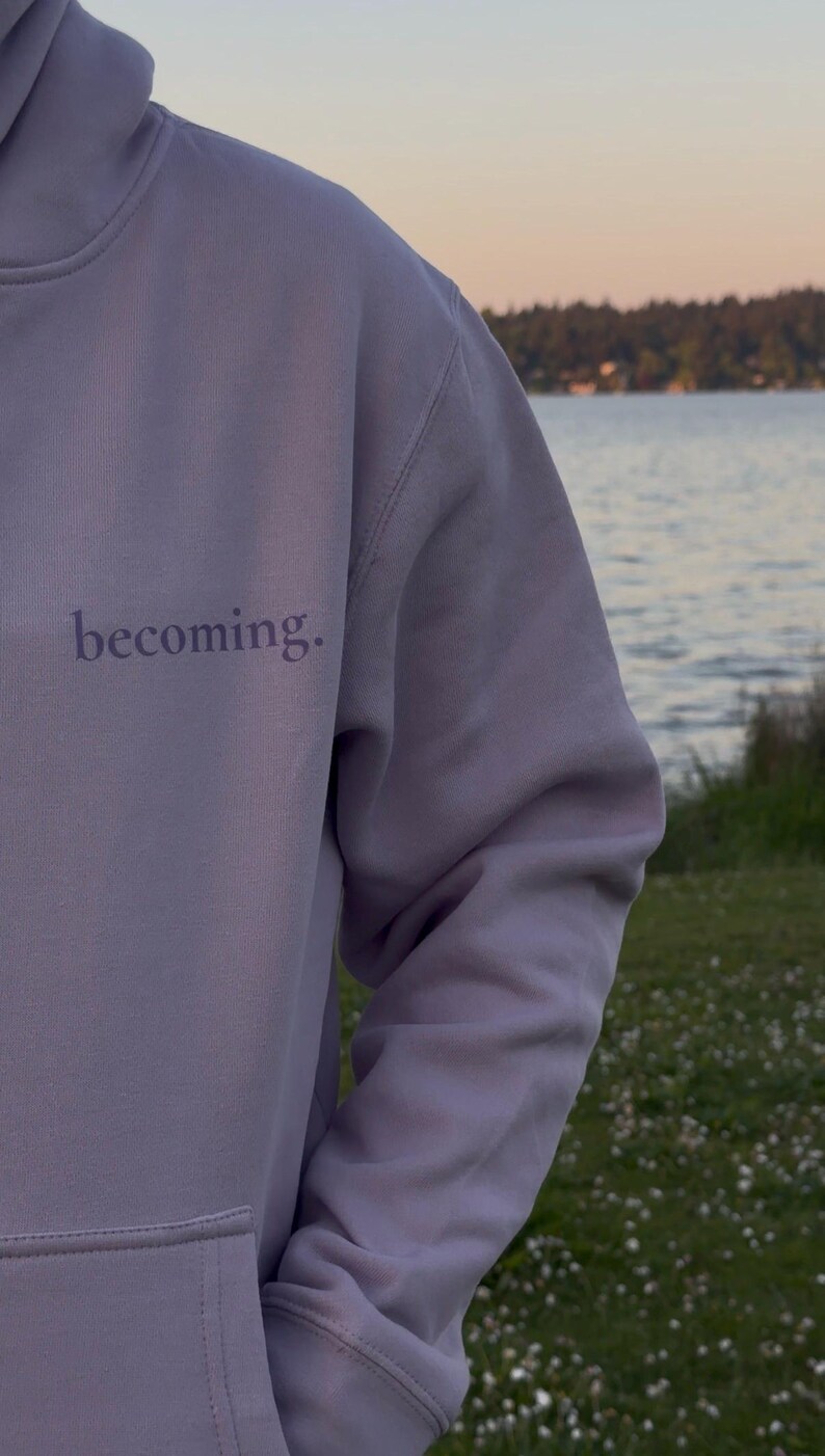 May include: A lavender hoodie with the word "becoming" printed on the front. The hoodie has a drawstring hood and a kangaroo pocket. The background shows a body of water and a treeline.