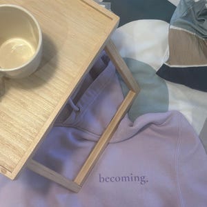 May include: A light purple hoodie with the word "becoming" printed on the front. A light brown wooden tray with a white mug is on top of the hoodie. The background is a patterned bedspread.