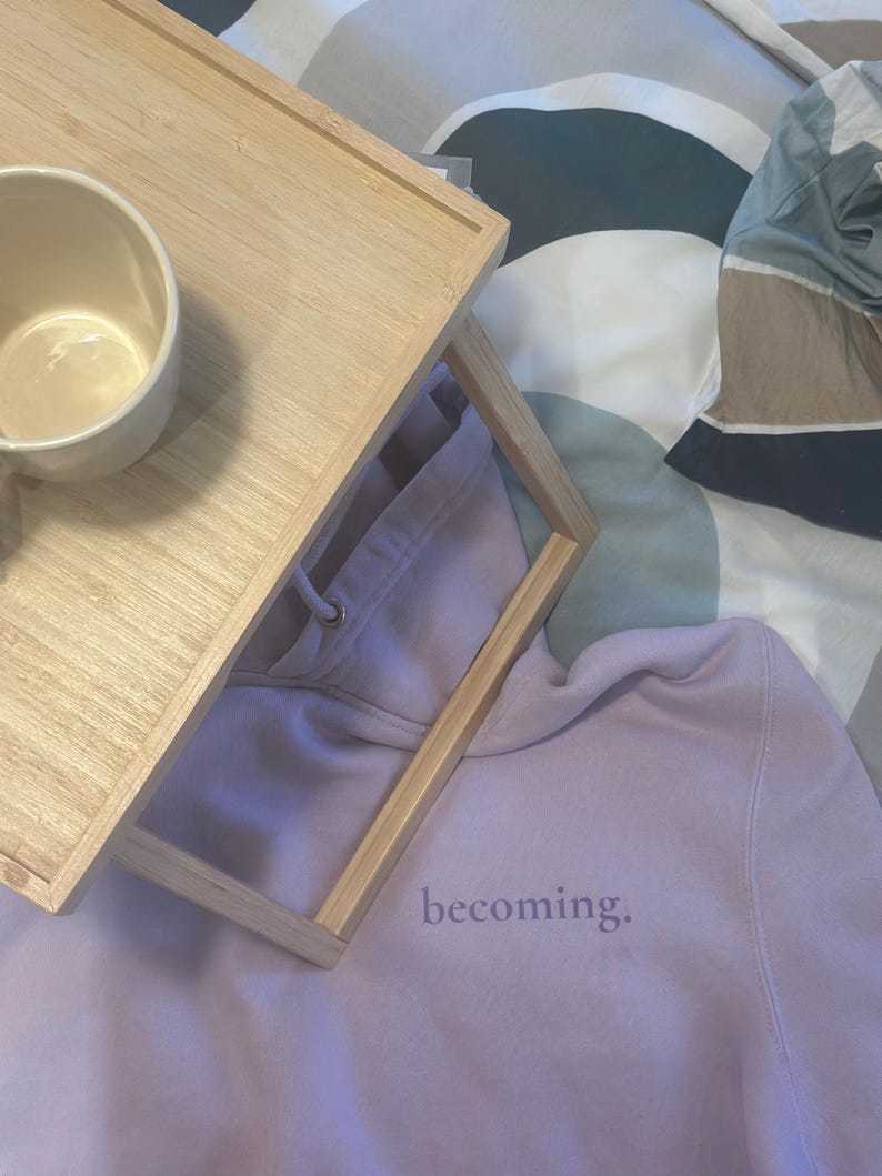 May include: A light purple hoodie with the word "becoming" printed on the front. A light brown wooden tray with a white mug is on top of the hoodie. The background is a patterned bedspread.