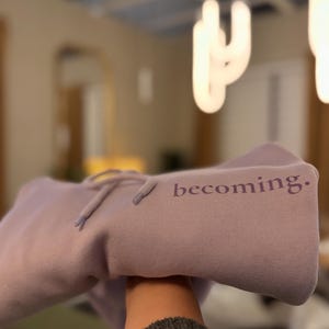 May include: A lavender hoodie with the word "becoming." printed on the front. The hoodie has a drawstring and is made of a soft material. The background is blurred, with a light fixture visible.