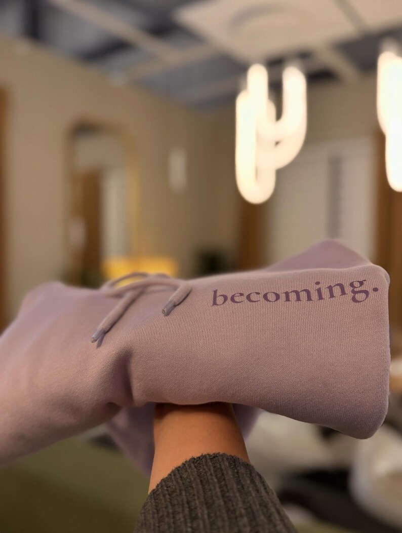 May include: A lavender hoodie with the word "becoming." printed on the front. The hoodie has a drawstring and is made of a soft material. The background is blurred, with a light fixture visible.