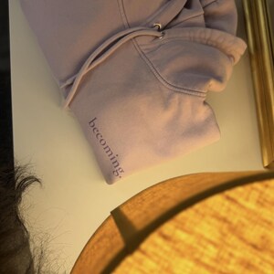 May include: A light purple hoodie with a drawstring hood lies on a white surface. The word "becoming" is printed in a darker purple on the sleeve. The hoodie is made of a soft material.