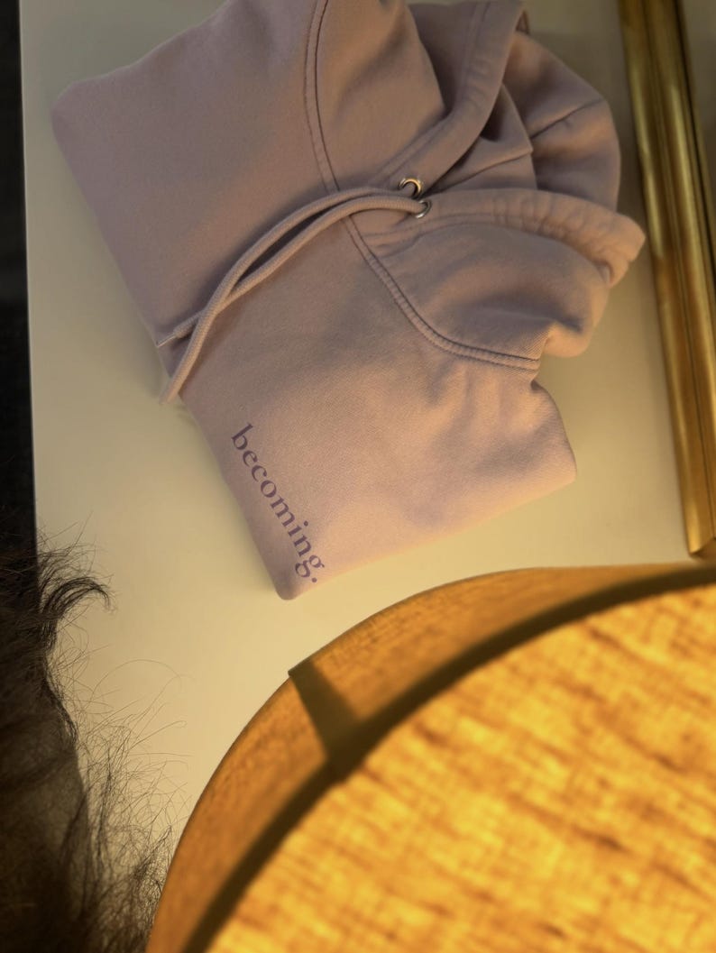 May include: A light purple hoodie with a drawstring hood lies on a white surface. The word "becoming" is printed in a darker purple on the sleeve. The hoodie is made of a soft material.