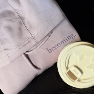 May include: A light purple hoodie with the word "becoming" printed on the front. The hoodie has a drawstring and a hood. A white coffee cup lid is in the foreground.