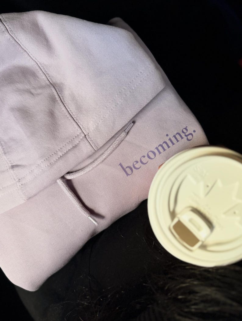 May include: A light purple hoodie with the word "becoming" printed on the front. The hoodie has a drawstring and a hood. A white coffee cup lid is in the foreground.