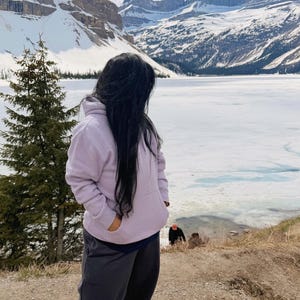 May include: A person wearing a light purple hoodie and dark gray sweatpants stands on a dirt path, looking towards a snow-covered mountain range and a partially frozen lake. The person is wearing brown hiking boots.
