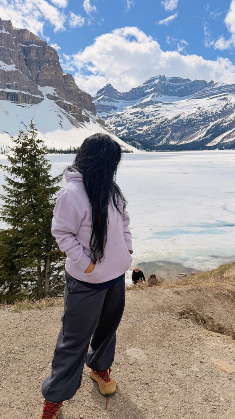 May include: A person wearing a light purple hoodie and dark gray sweatpants stands on a dirt path, looking towards a snow-covered mountain range and a partially frozen lake. The person is wearing brown hiking boots.