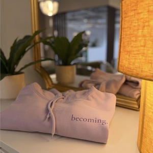 May include: A folded lavender hoodie with the word "becoming" embroidered on the front. The hoodie has a drawstring and is resting on a white surface. A gold-framed mirror and a lit lamp are in the background.