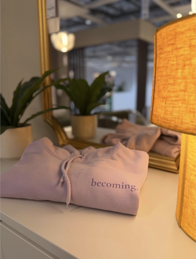 May include: A folded lavender hoodie with the word "becoming" embroidered on the front. The hoodie has a drawstring and is resting on a white surface. A gold-framed mirror and a lit lamp are in the background.