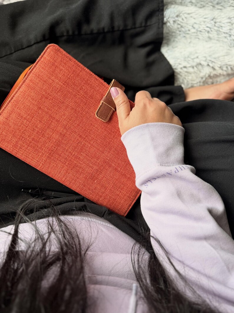 May include: An orange fabric-covered journal or notebook with a brown leather strap closure. The journal is rectangular and appears to be closed. The background is dark, with a person's arm and clothing visible.