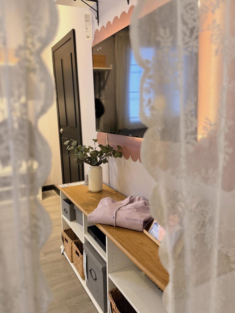 May include: A light wood entertainment center with open shelves and storage baskets. A lavender hoodie with the word "becoming" is on top. A black TV is mounted on the wall above the center.