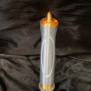 K&ouml;nnte beinhalten: Ein silberner und goldener Lichtschwert-Requisit mit einer spitzen Spitze und einem strukturierten Griff.