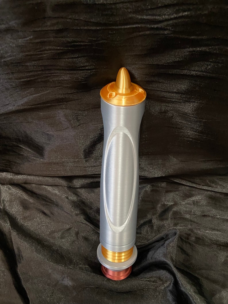 K&ouml;nnte beinhalten: Ein silberner und goldener Lichtschwert-Requisit mit einer spitzen Spitze und einem strukturierten Griff.