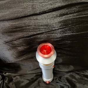 K&ouml;nnte beinhalten: Ein wei&szlig;es Lichtschwert mit einem roten, kupfernen und goldenen Griff. Das Lichtschwert liegt auf einem schwarzen Stoffhintergrund.